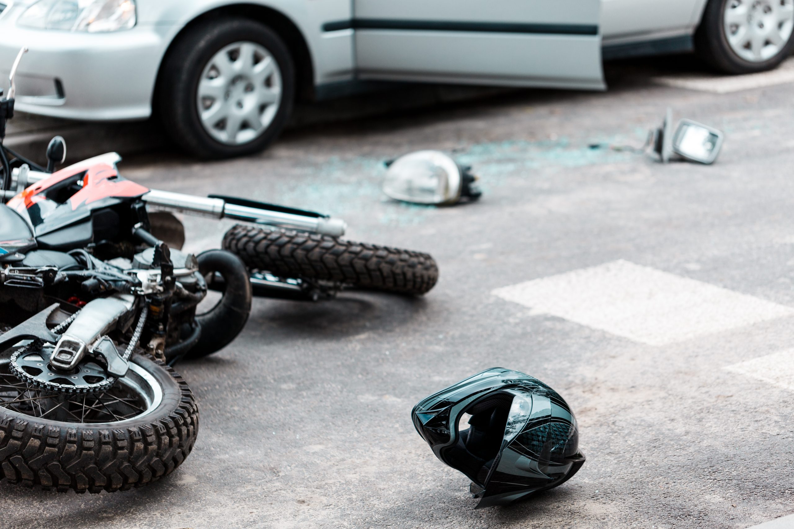 Wrecked motorcycle on pavement with broken parts, a helmet nearby, and a car in the background after an accident
