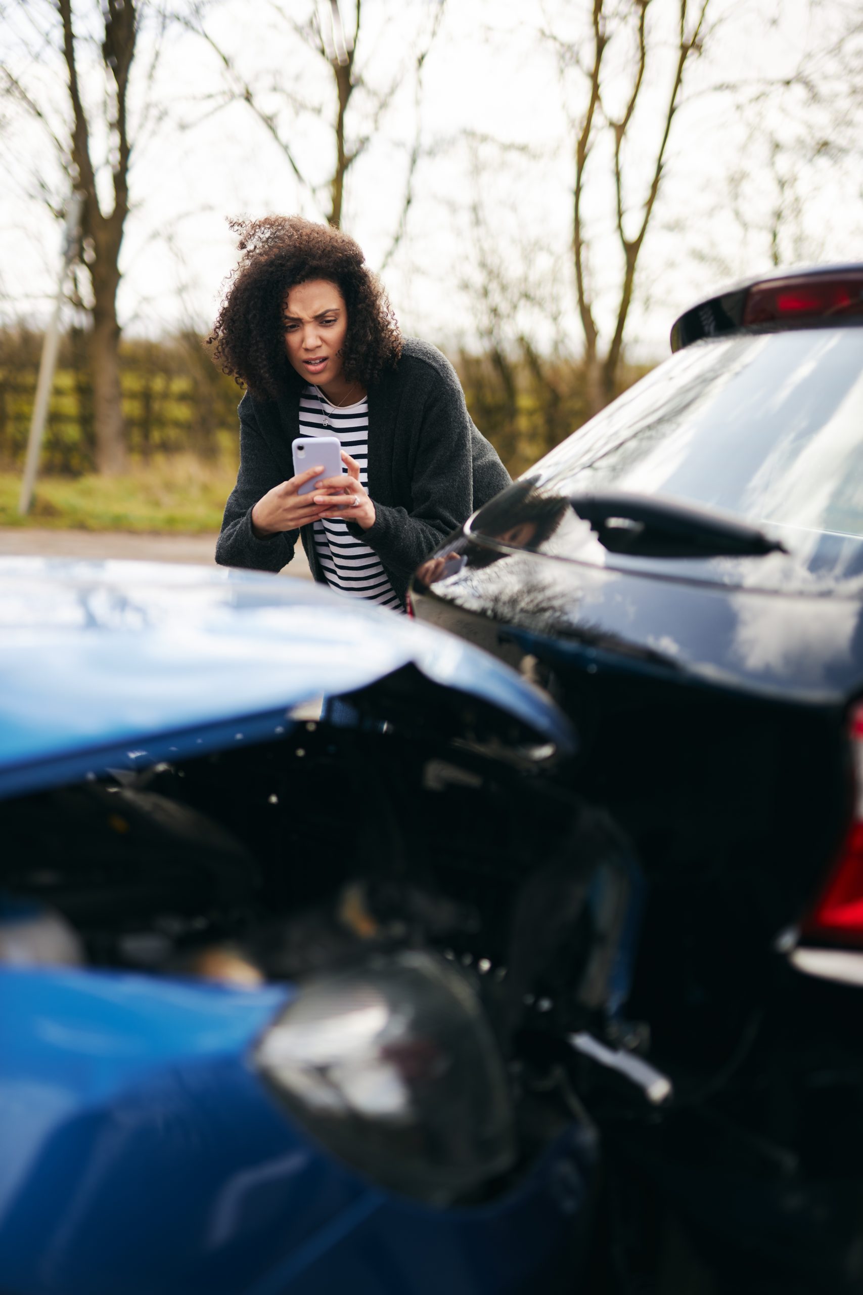 Woman with curly hair looking at her phone while standing between two collided cars in a parking lot, outdoors with bare trees in the background.